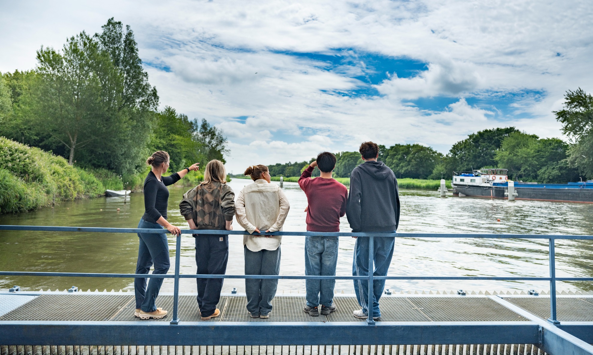 Studenten en docent staan langs het water en wijzen naar de langsvarende boot.