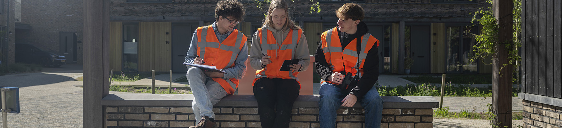 Drie studenten gekleed in oranje veiligheidshesjes op een muurtje.